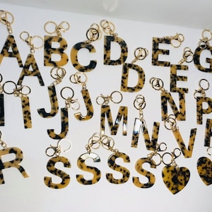 May include: A set of 18 gold keychains with letter charms. The charms are made of a brown and black mottled material, resembling tortoiseshell. The letters include A, B, C, D, E, F, G, H, I, J, K, L, M, N, O, P, Q, R, S, T, U, V, W, X, Y, and Z.