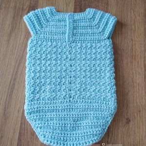May include: A light blue crocheted baby onesie with short sleeves. The onesie has a textured stitch pattern and a button closure in the back.