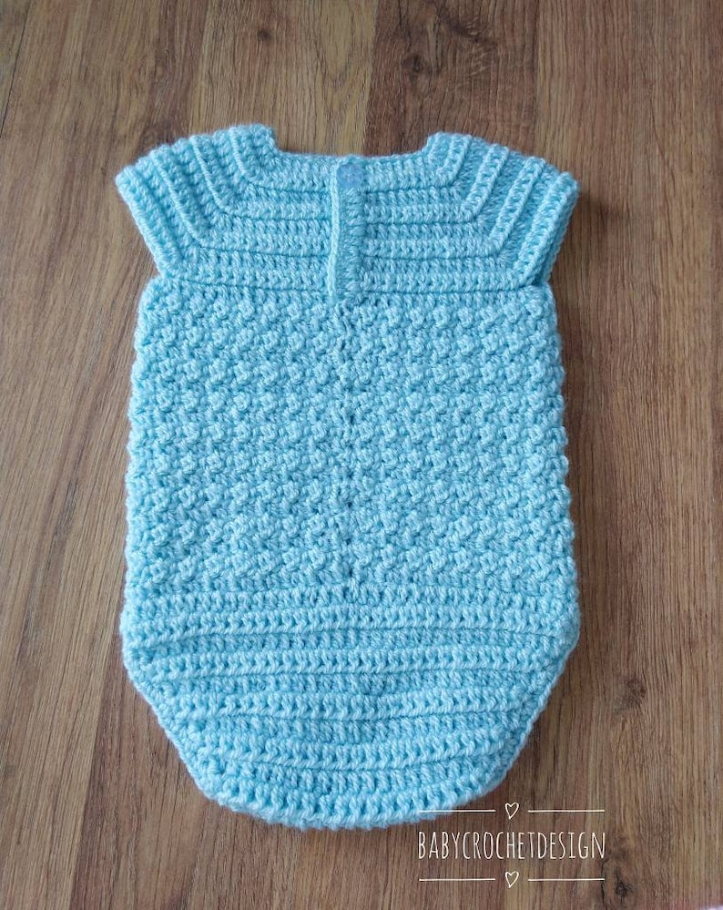 May include: A light blue crocheted baby onesie with short sleeves. The onesie has a textured stitch pattern and a button closure in the back.