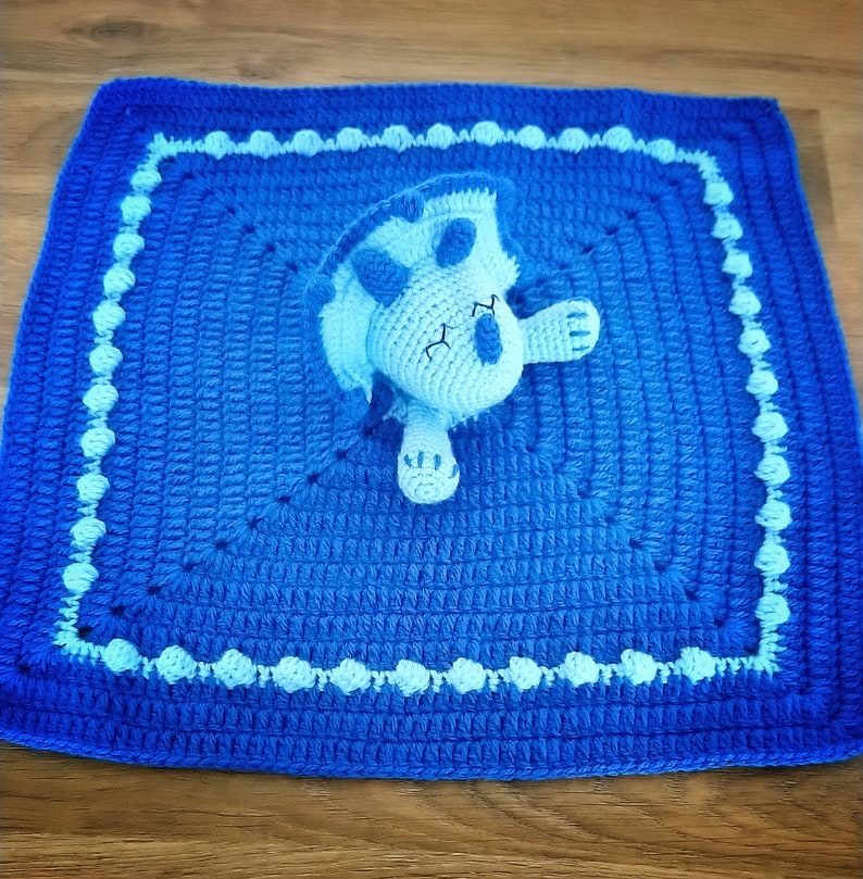 May include: A blue crocheted baby blanket with a light blue crocheted dinosaur attached. The blanket has a white border with small white circles.