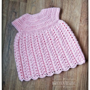 May include: A pink crocheted baby dress with a scalloped hem and a round neckline. The dress is made with a delicate stitch pattern and has short sleeves.