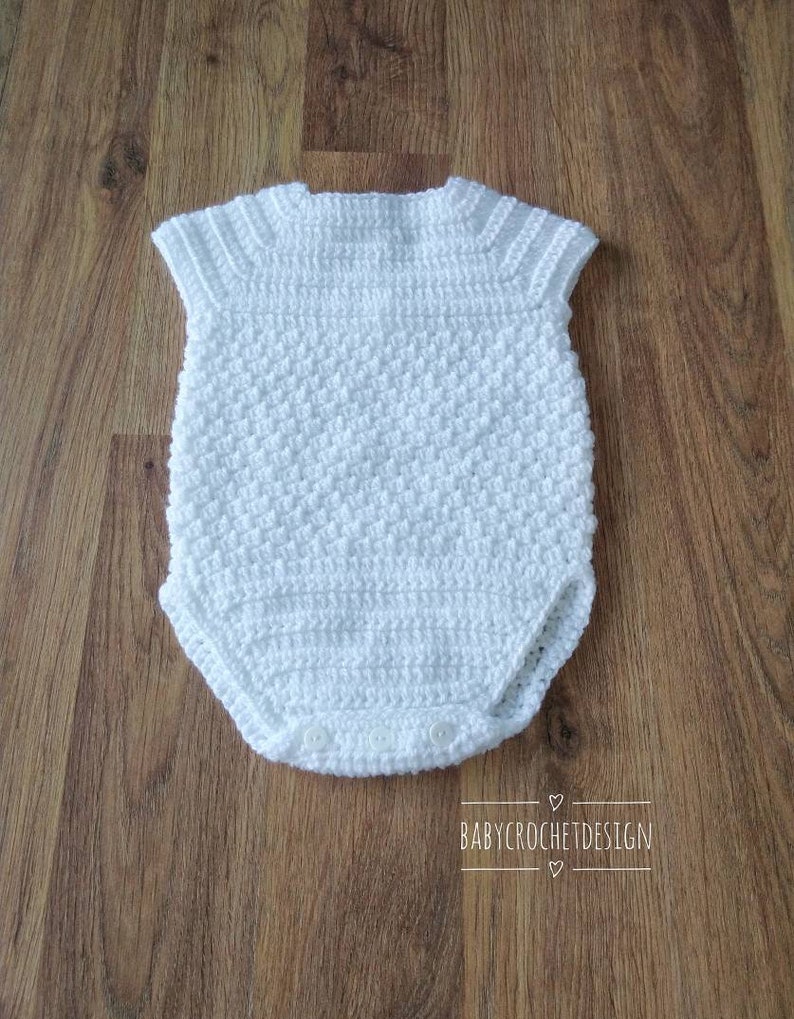 May include: White crocheted baby onesie with short sleeves and a button closure. The onesie is made with a textured stitch and has a simple design.