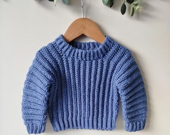 May include: A blue crocheted sweater for a baby or toddler. The sweater has a ribbed texture and long sleeves. The sweater is hanging on a wooden hanger against a white background.