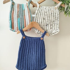 May include: Three handmade crochet baby rompers in the image. One is blue, one is striped with blue, peach, and white, and the other is speckled white. Each has wooden button closures and hangs on a wooden hanger. The website address is visible at the bottom.
