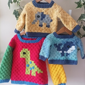 May include: Three handmade crochet sweaters for children. The sweaters are yellow, red, and blue, each with an animal design: an elephant, a dinosaur, and a whale. The sweaters have blue trim and are displayed on wooden hangers.