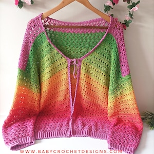 Puede incluir: Un cárdigan de ganchillo hecho a mano, de color arcoíris, con cuello en V y mangas largas. El degradado de color va del rosa al verde y al naranja. El cárdigan se muestra en una percha de madera. La dirección del sitio web www.babycrochetdesigns.com es visible.