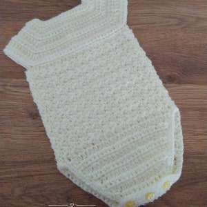 May include: A white crocheted baby onesie with yellow buttons. The onesie has a simple design and is perfect for a newborn.