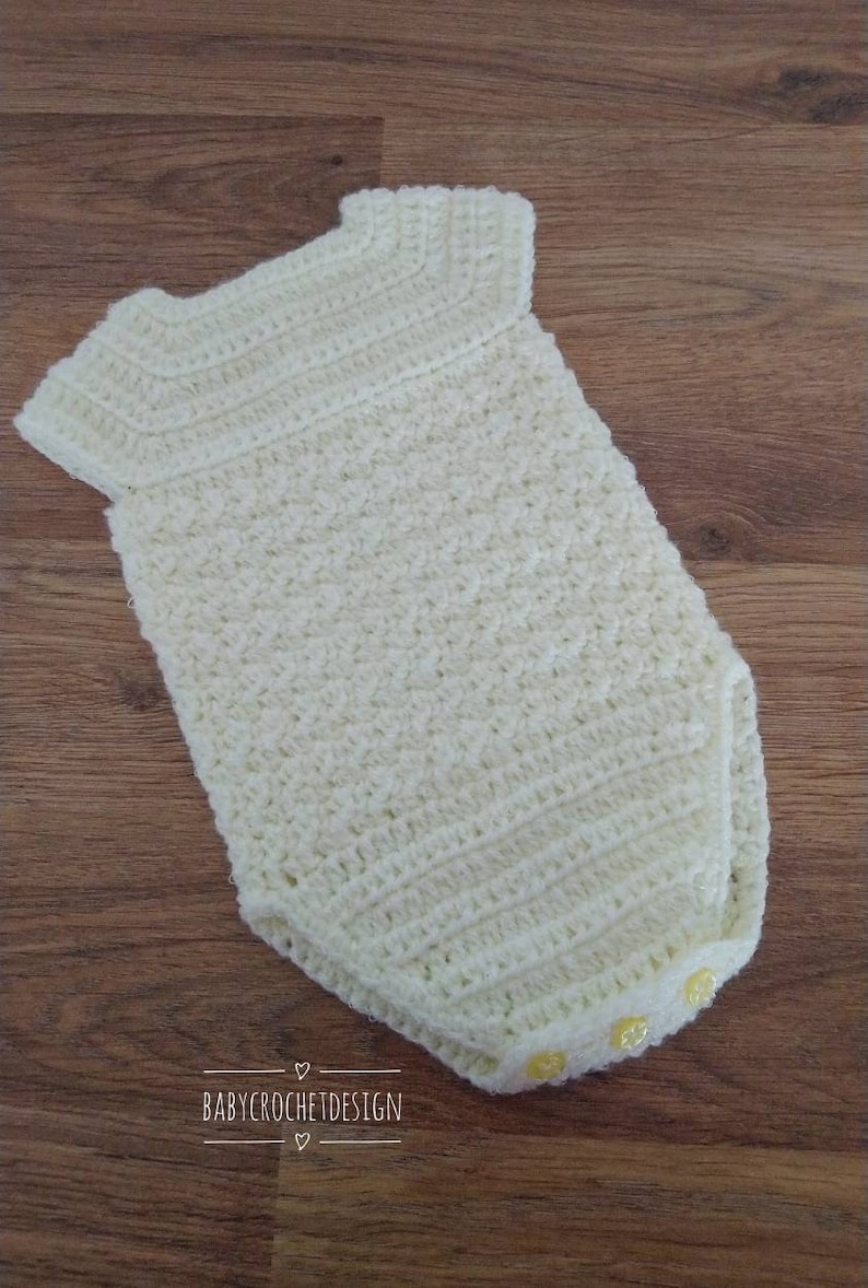 May include: A white crocheted baby onesie with yellow buttons. The onesie has a simple design and is perfect for a newborn.