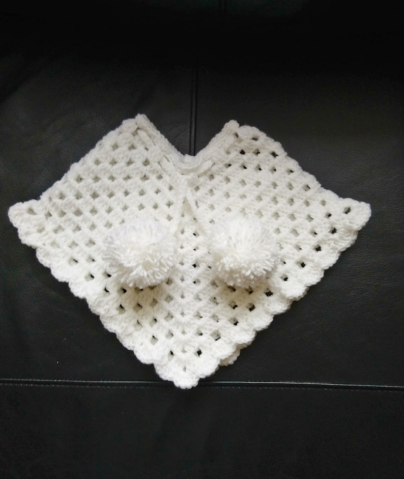 K&ouml;nnte beinhalten: Wei&szlig;es H&auml;kelponcho mit zwei wei&szlig;en Bommeln.