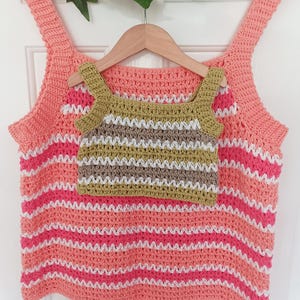 May include: A crocheted tank top with a striped pattern in shades of pink, white, and coral. A smaller crocheted tank top with a striped pattern in shades of green, white, and brown is layered on top. The tops are hanging on a wooden hanger.