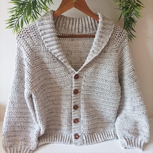 May include: A light gray, hand-crocheted cardigan with a shawl collar and wooden buttons. The long-sleeved sweater is displayed on a wooden hanger. The website address www.babycrochetdesigns.com is visible at the bottom of the image.
