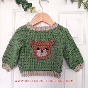 Könnte beinhalten: Ein grüner Häkelpullover für Babys mit einem braunen Bärenmotiv auf der Vorderseite. Der Pullover hat einen Rundhalsausschnitt und gerippte Bündchen und Saum in einer neutralen Farbe. Die Website-Adresse www.babycrochetdesigns.com ist unten aufgedruckt.