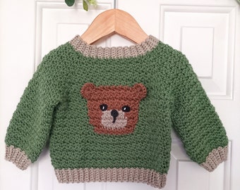 May include: A green crochet baby sweater with a brown bear design on the front. The sweater has a crew neck and ribbed cuffs and hem in a neutral colour. The website address www.babycrochetdesigns.com is printed at the bottom.