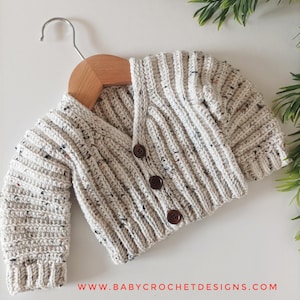 May include: A cream-coloured crocheted baby cardigan with brown buttons. The cardigan is hanging on a wooden hanger. The text "www.babycrochetdesigns.com" is visible at the bottom of the image.