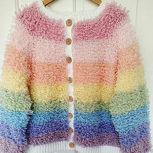 May include: A colourful, handmade crochet cardigan with a fluffy texture. The sweater has a round neckline, wooden buttons, and a white trim. The rainbow colour scheme includes pink, orange, yellow, green, blue, and purple. The website address is visible at the bottom.