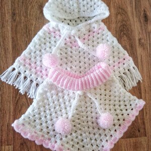 K&ouml;nnte beinhalten: Wei&szlig;es geh&auml;keltes Kapuzenponcho mit rosa Besatz und Bommeln. Das Poncho hat einen Fransenrand und der Text "BABY CROCHET DESIGNS" ist am unteren Rand des Bildes sichtbar.