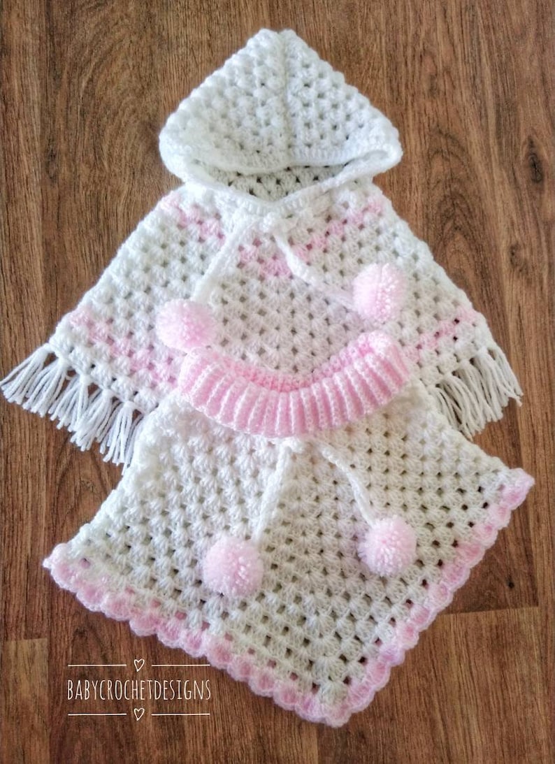 K&ouml;nnte beinhalten: Wei&szlig;es geh&auml;keltes Kapuzenponcho mit rosa Besatz und Bommeln. Das Poncho hat einen Fransenrand und der Text "BABY CROCHET DESIGNS" ist am unteren Rand des Bildes sichtbar.