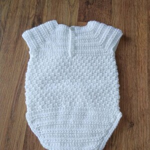 May include: White crocheted baby onesie with a button closure at the back. The onesie has a textured stitch pattern and short sleeves.