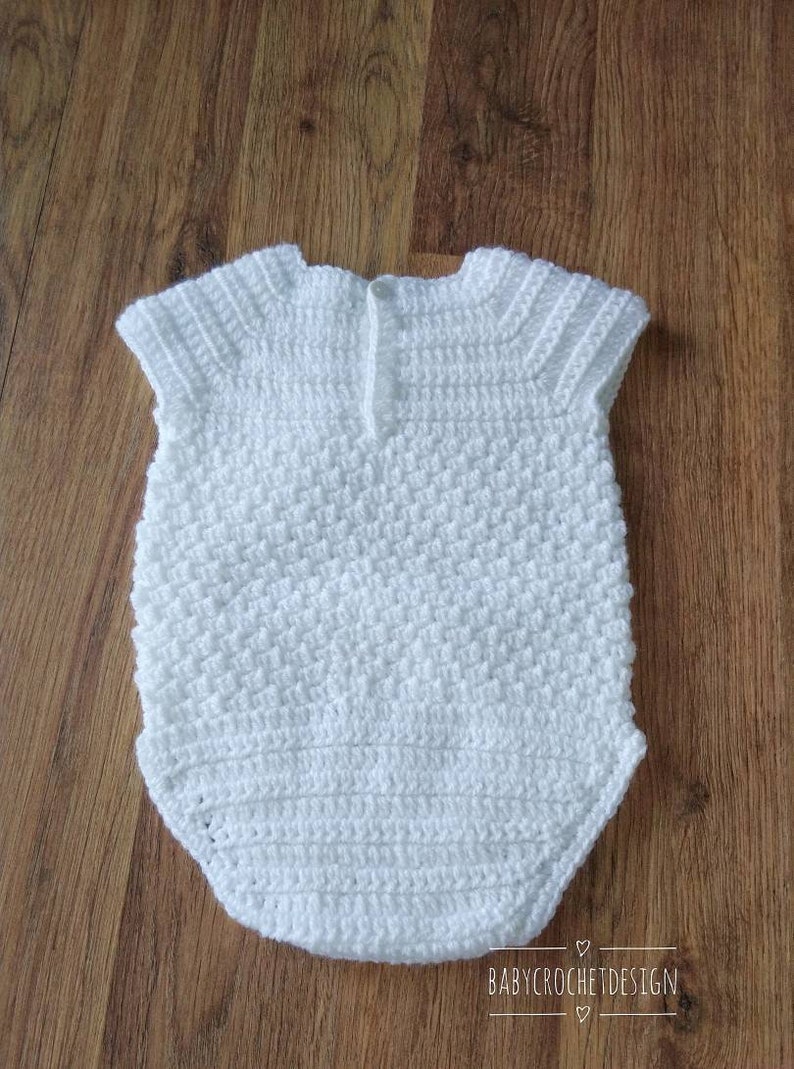 May include: White crocheted baby onesie with a button closure at the back. The onesie has a textured stitch pattern and short sleeves.