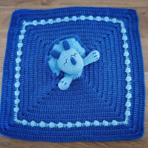 May include: A blue crocheted baby blanket with a light blue crocheted dinosaur in the centre.