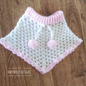 K&ouml;nnte beinhalten: Wei&szlig;es H&auml;kel-Babyponcho mit rosa Besatz und Bommeln. Das Poncho hat einen gewellten Rand und einen rosa Streifen um den Hals. Der Text "BABY CROCHET DESIGNS" ist am unteren Rand des Bildes sichtbar.