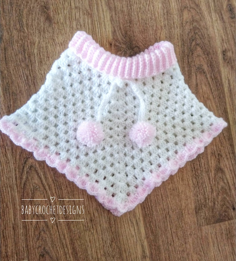 K&ouml;nnte beinhalten: Wei&szlig;es H&auml;kel-Babyponcho mit rosa Besatz und Bommeln. Das Poncho hat einen gewellten Rand und einen rosa Streifen um den Hals. Der Text "BABY CROCHET DESIGNS" ist am unteren Rand des Bildes sichtbar.