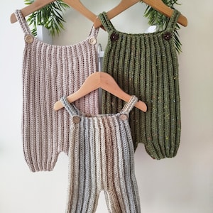 Puede incluir: Tres peleles de bebé hechos a mano con ganchillo en colores neutros cuelgan de perchas de madera. Los peleles son en tonos rosa, verde y gris, con cierres de botones de madera. La textura acanalada y el diseño sencillo crean una estética acogedora y rústica.