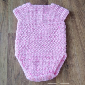 May include: A pink crocheted baby onesie with short sleeves and button closures. The onesie is made with a textured stitch and has a subtle sparkle.