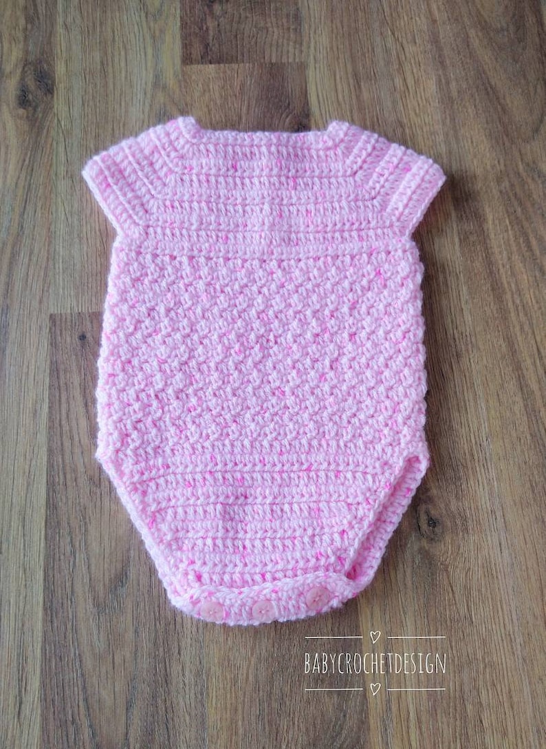 May include: A pink crocheted baby onesie with short sleeves and button closures. The onesie is made with a textured stitch and has a subtle sparkle.