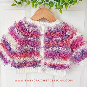 May include: A white crocheted cardigan with pink, purple and white yarn. The cardigan has a button fastening and is hanging on a wooden hanger.