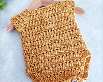 May include: A crocheted baby onesie in a mustard yellow color with a delicate open stitch pattern. The onesie has three wooden buttons on the bottom.