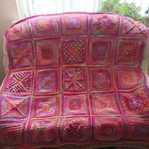 May include: A handmade crochet blanket in shades of pink, red, and orange. The blanket is composed of numerous square panels, each with a unique textured design. The overall pattern is a patchwork of intricate crochet stitches.
