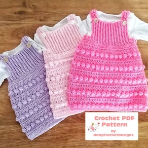 May include: Three crocheted baby dresses in purple, pink, and white with a crocheted bobble stitch. The dresses have straps and buttons. The dresses are on a wooden surface. Crochet PDF Pattern by BabyCrochetDesigns.