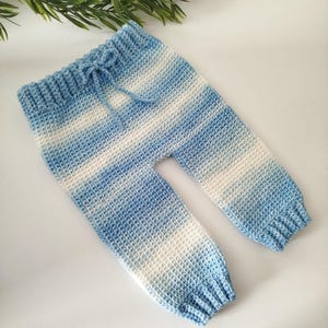 May include: Crocheted baby pants in shades of blue and white. The pants have a drawstring waist and ribbed cuffs. The yarn creates a striped effect. The website address www.babycrochetdesigns.com is printed at the bottom.