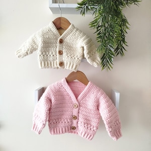May include: Two baby cardigans, one cream and one pink, hang on wooden hangers. The cream cardigan features a textured stitch and wooden buttons. The pink cardigan has a simpler stitch and wooden buttons. Both are displayed against a neutral background.