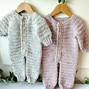 May include: Two crocheted baby onesies, one in a light green color and the other in a light pink color, hanging on wooden hangers. Both onesies have wooden buttons down the front.