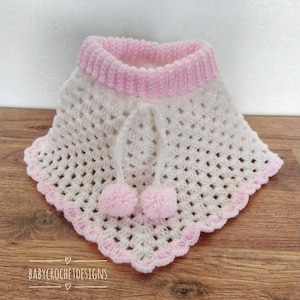 K&ouml;nnte beinhalten: Wei&szlig;es geh&auml;keltes Babyponcho mit rosa Besatz und zwei rosa Bommeln. Das Poncho hat eine wei&szlig;e geh&auml;kelte Kapuze mit einem rosa geh&auml;kelten Band.