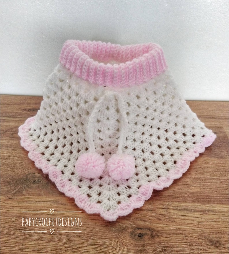 K&ouml;nnte beinhalten: Wei&szlig;es geh&auml;keltes Babyponcho mit rosa Besatz und zwei rosa Bommeln. Das Poncho hat eine wei&szlig;e geh&auml;kelte Kapuze mit einem rosa geh&auml;kelten Band.