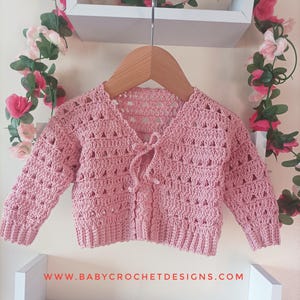 May include: A pale pink, open-weave crochet baby cardigan with long sleeves, hanging on a wooden hanger. The cardigan has a tie closure at the neckline. The website address www.babycrochetdesigns.com is visible at the bottom.