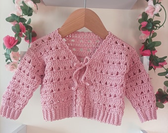 May include: A light pink, open-weave crochet baby cardigan with long sleeves, hanging on a wooden hanger. The cardigan has a tie closure at the neckline. The website address www.babycrochetdesigns.com is visible at the bottom.