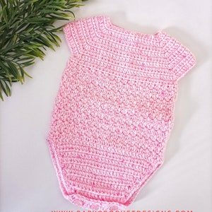 May include: A pink crocheted baby onesie with short sleeves. The onesie has a simple design and is made with a soft yarn.