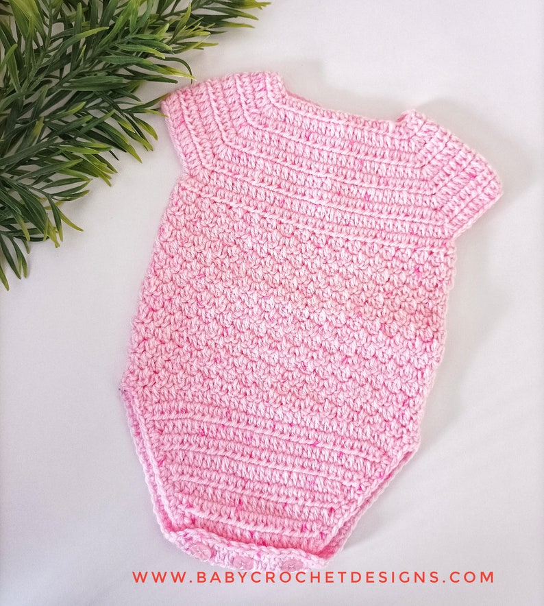 May include: A pink crocheted baby onesie with short sleeves. The onesie has a simple design and is made with a soft yarn.