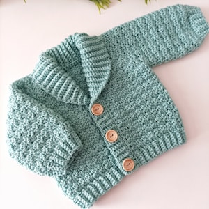 May include: A light green crocheted baby cardigan with a collar and wooden buttons. The cardigan is laid flat on a white surface.
