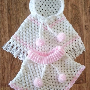 K&ouml;nnte beinhalten: Wei&szlig;es geh&auml;keltes Babyponcho mit rosa Besatz und Bommeln. Das Poncho hat eine Kapuze und Fransen.