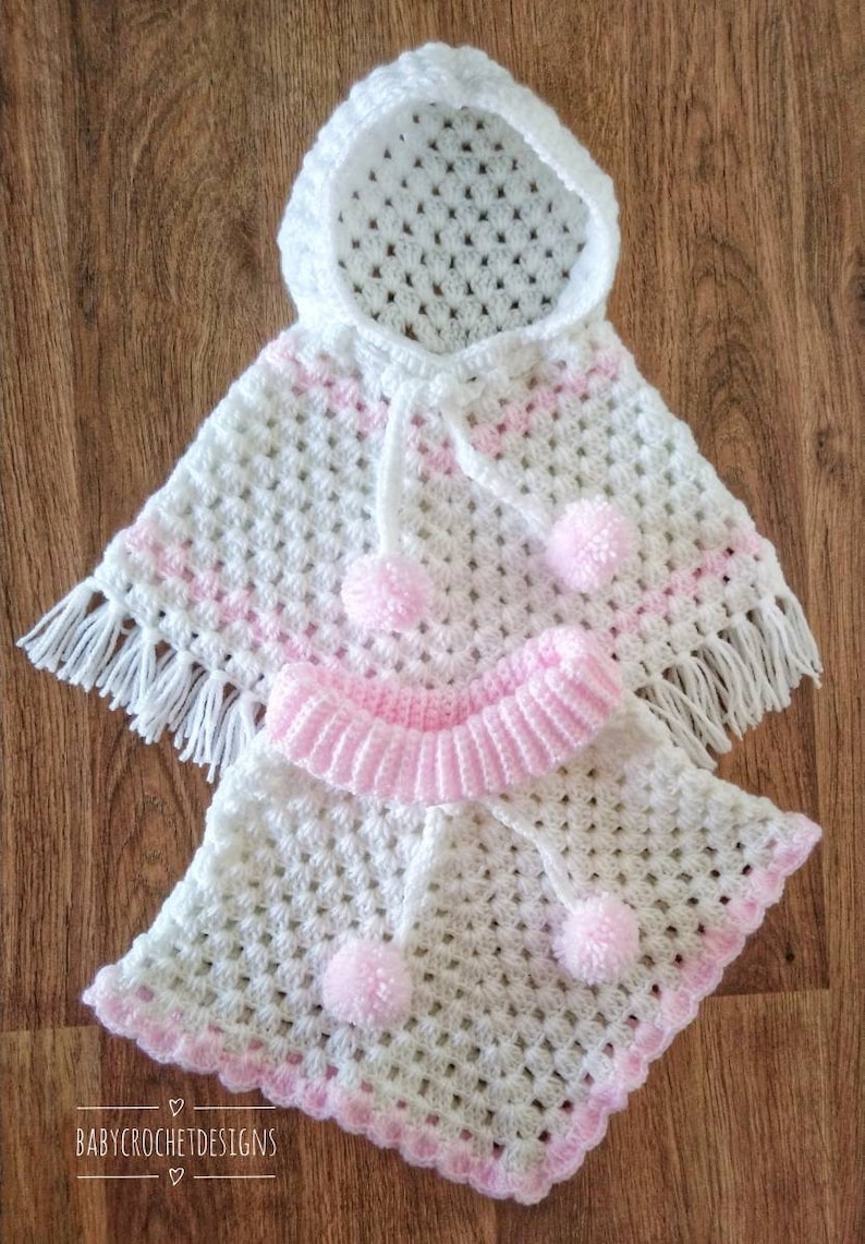 K&ouml;nnte beinhalten: Wei&szlig;es geh&auml;keltes Babyponcho mit rosa Besatz und Bommeln. Das Poncho hat eine Kapuze und Fransen.