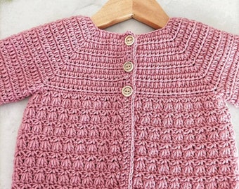 May include: A pink crocheted baby cardigan with button closures. The cardigan is hanging on a wooden hanger. Crochet PDF Pattern by BabyCrochetDesigns.