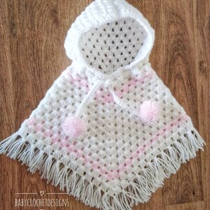 K&ouml;nnte beinhalten: Wei&szlig;es geh&auml;keltes Kapuzenponcho mit rosa Streifen und wei&szlig;em Fransenbesatz. Das Poncho hat zwei wei&szlig;e Bommeln am unteren Rand.