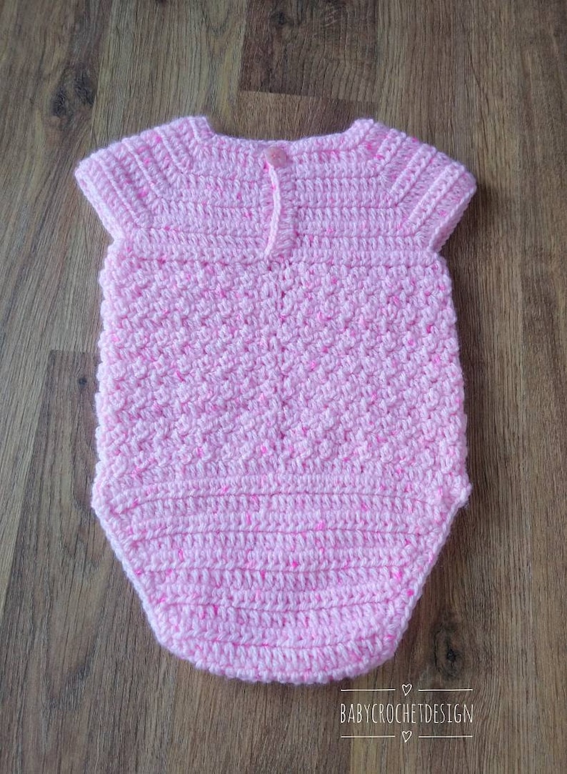 May include: A pink crocheted baby onesie with short sleeves. The onesie has a button closure at the back.
