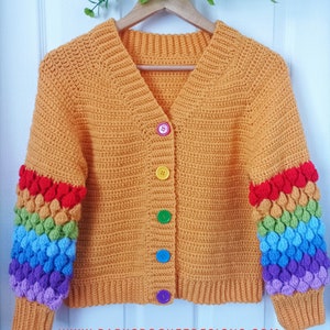 May include: A crocheted cardigan with a yellow body and rainbow colored sleeves. The sleeves have a textured design and the cardigan has a v-neck and buttons down the front. The buttons are multi-colored. The cardigan is hanging on a wooden hanger. The text "www.babycrochetdesigns.com" is at the bottom of the image.