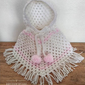 K&ouml;nnte beinhalten: Wei&szlig;es geh&auml;keltes Kapuzenponcho mit rosa Streifen und wei&szlig;en Fransen. Das Poncho hat zwei rosa Bommeln, die an der Kapuze befestigt sind.